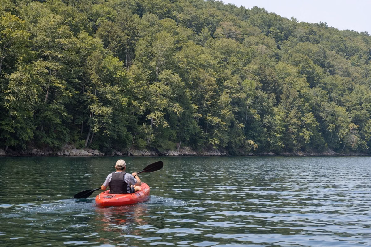 A kayaker paddling on calm blue-green water near a wooded shore